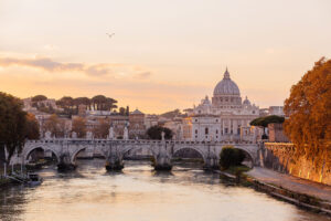 A Roman stone bridge spans the Tiber River at sunset.