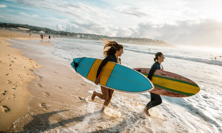 Two young women run into the North Coast surf with surfboards.