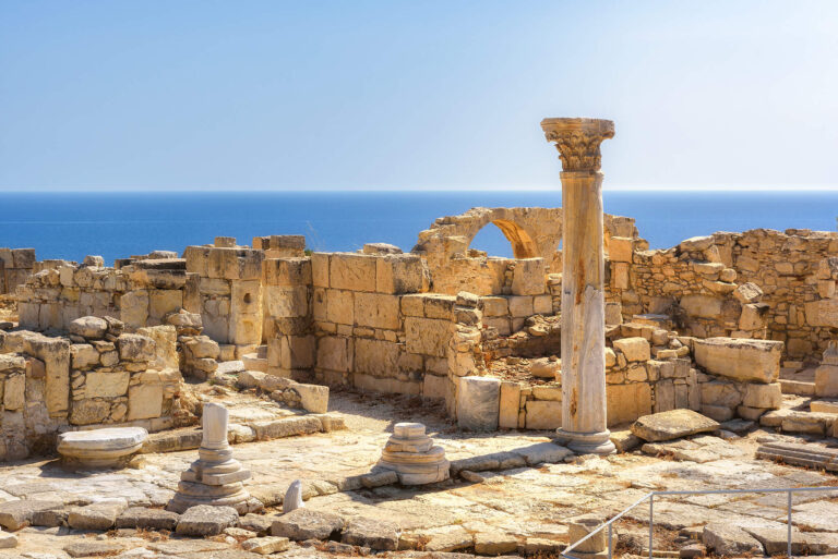 Old pillars and stone ruins of the ancient Kingdom of Kourion overlook a hazy blue ocean.