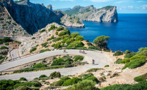 Bikers pedal uphill on a road paved through the rocky Mallorca coastline.