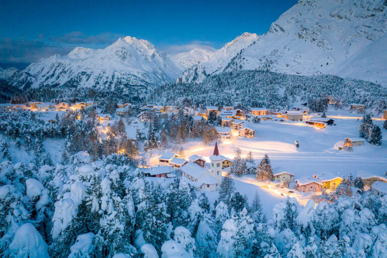 Idyllic Swiss village with gentle yellow lights surrounded by alpine mountains and covered with snow.