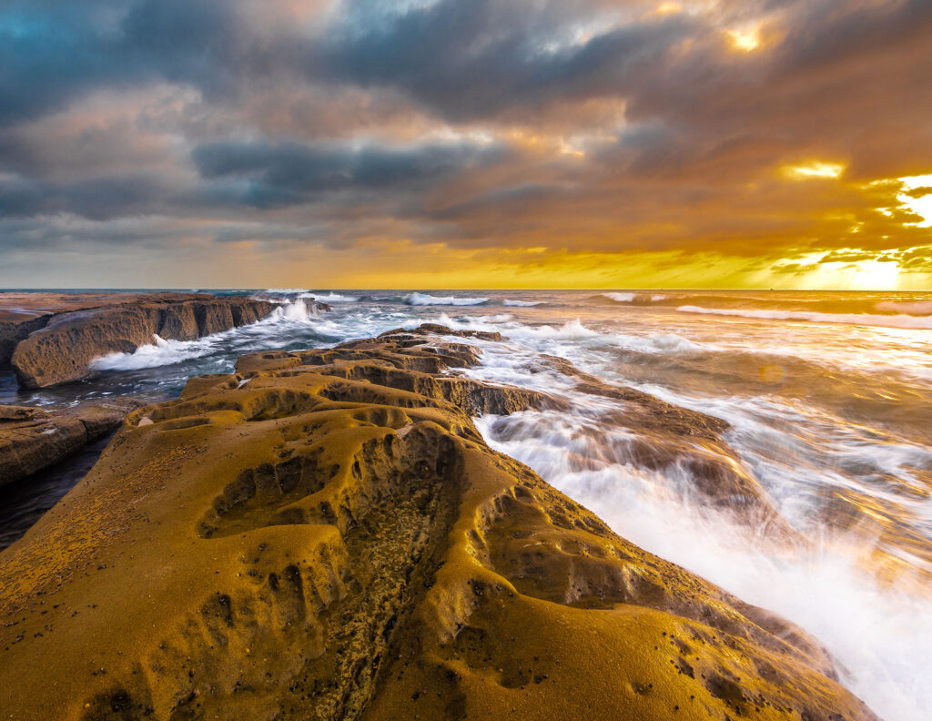 The sea splashes over surf-eroded rocks against a cloudy California sky.
