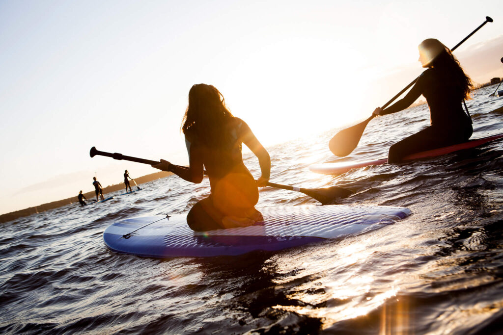 Two young women paddleboarding in wetsuits look to the sunrise.