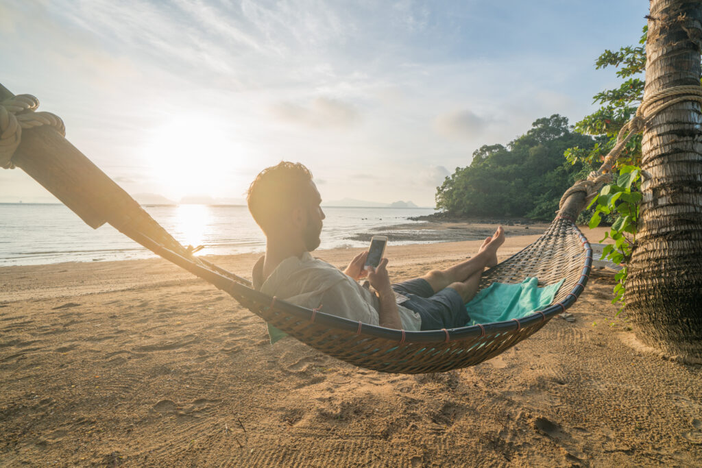 Man in shorts relaxes in a beachside hammock while browsing his mobile phone.