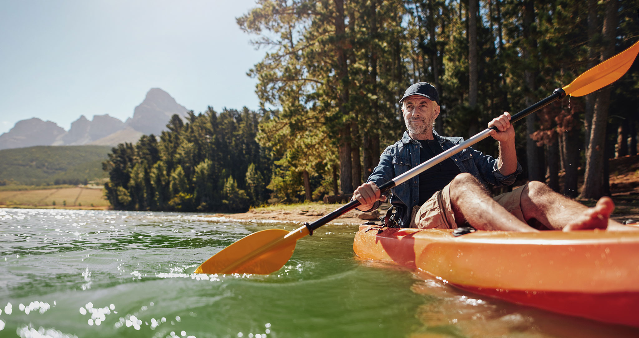 An older man in shorts paddles a kayak on a lake.