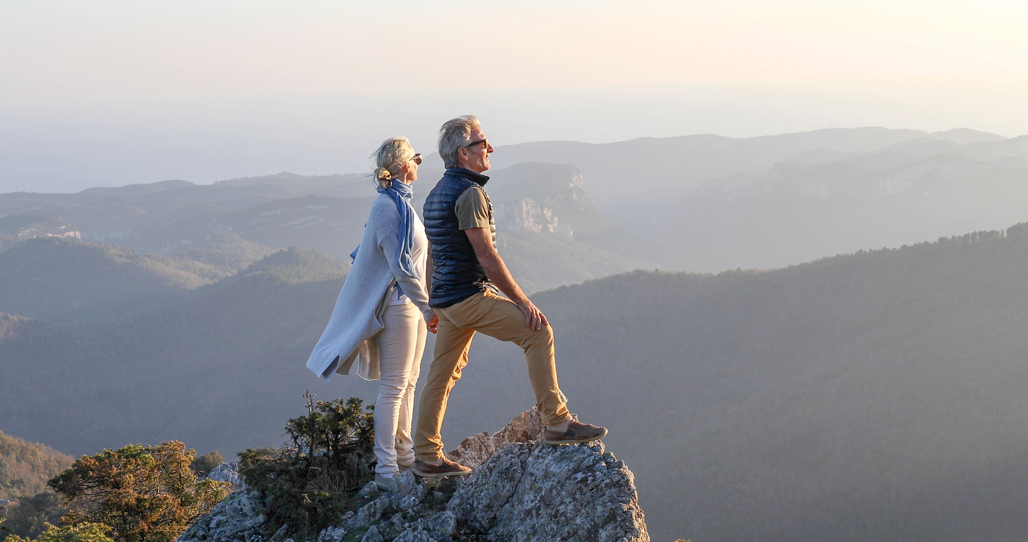 A senior couple stands on a rock overlooking distant mountains.