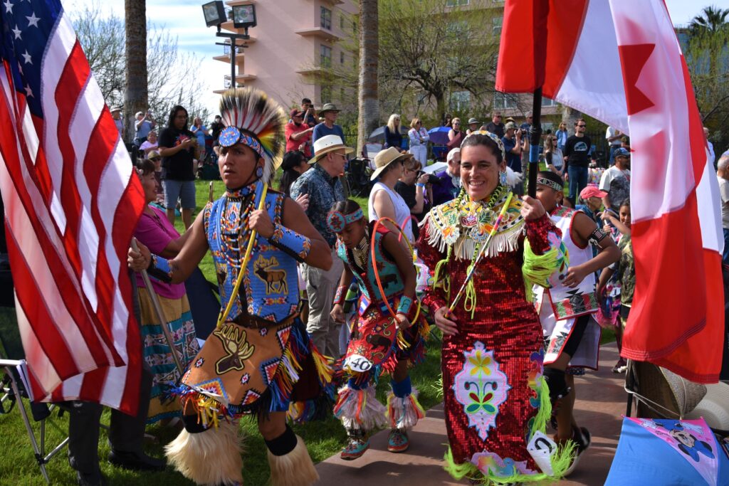 A vibrant group of Native American dancers dressed in traditional regalia participate in a cultural parade, carrying American and Canadian flags.
