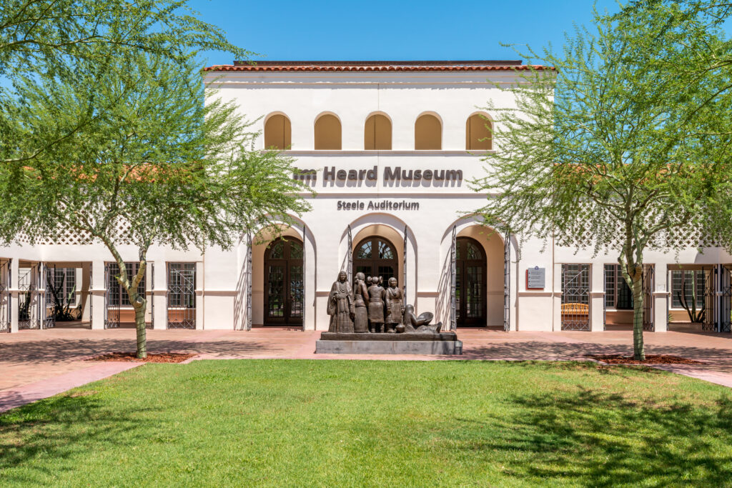 The front facade of the Heard Museum in Phoenix, Arizona, showcasing its Spanish Colonial Revival architecture with arched doorways and windows, surrounded by lush green trees.