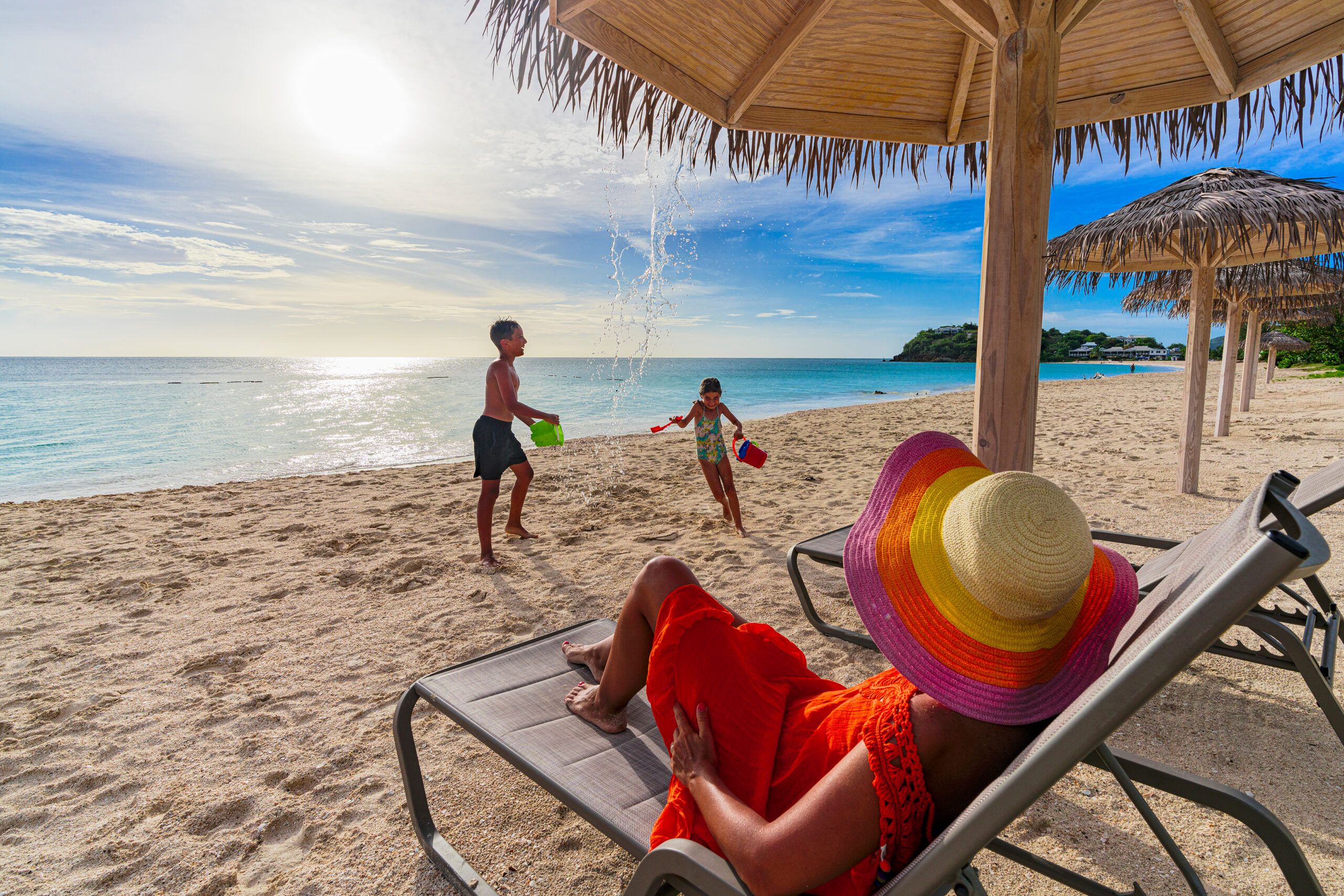 Woman on sun lounger looking at her happy kids playing on a beach at sunset, Antigua, Antigua & Barbuda, Caribbean
