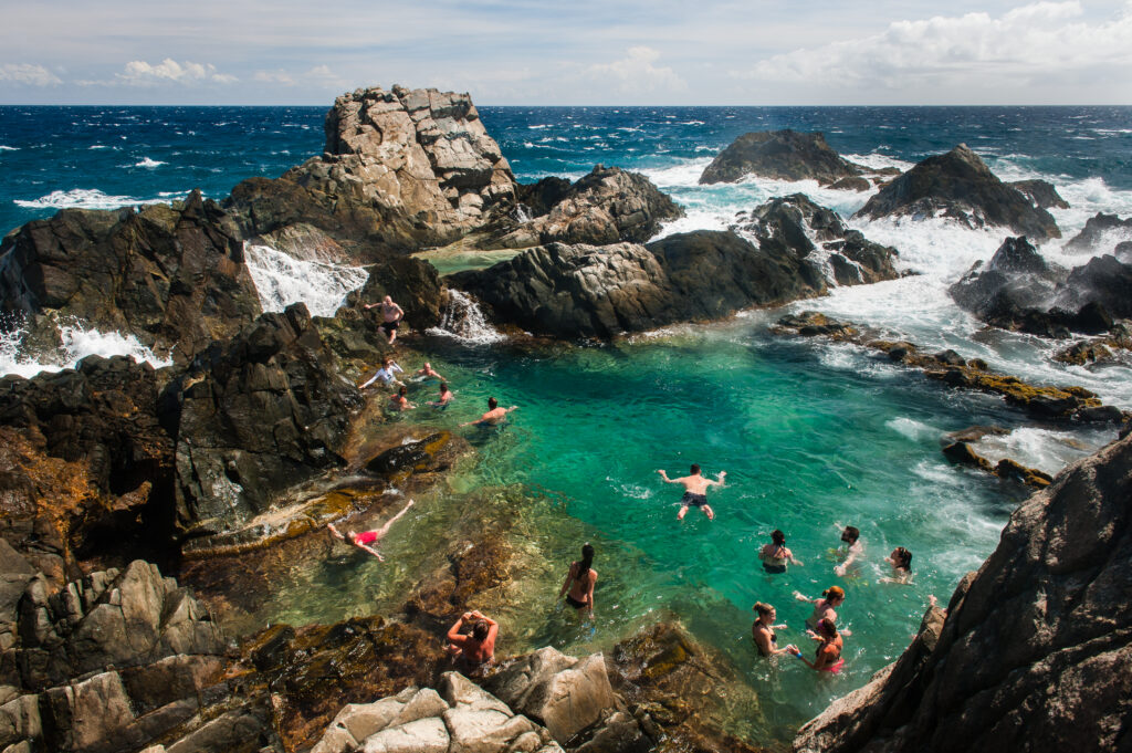 Visitors swimming in a natural turquoise rock pool surrounded by rugged coastal cliffs and ocean waves.