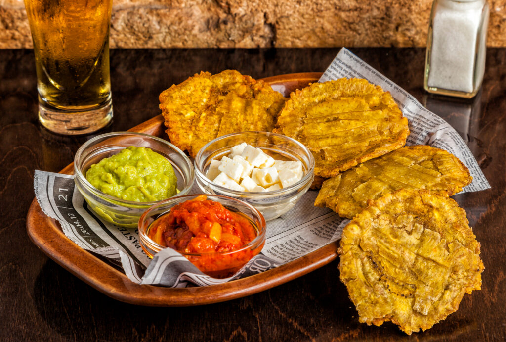 Traditional Caribbean tostones served with dipping sauces and cheese on a wooden platter alongside a cold beer.
