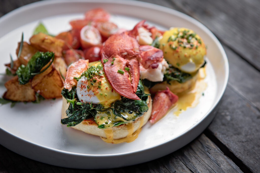 Lobster eggs Benedict served with sautéed greens, roasted potatoes, and tomatoes on a white plate.